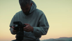 Photographer holding a professional video camera during golden hour with mountains visible in the background.