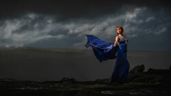 Woman in flowing blue gown standing on rocky shore with dramatic storm clouds overhead.