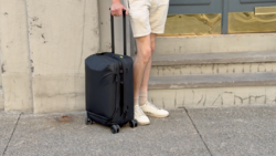 Person standing with black rolling carry-on luggage on concrete pavement beside a building.
