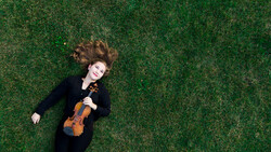 Musician lying on grass holding a violin, wearing dark clothing with hair spread across the ground.