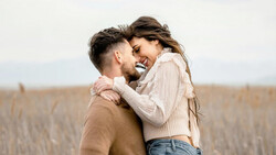Couple embracing and smiling in an open field with dried grass and overcast sky.