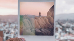 Polaroid photograph held in front of a blurred cityscape, showing a lone figure standing atop a large rock formation at sunset.
