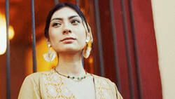 Woman wearing a patterned beige tunic and statement earrings gazes upward against warm amber and red draped backdrop.