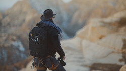 Hiker with backpack and camera gear standing on a mountain overlook facing rocky peaks.