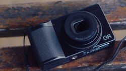 Ricoh GR compact camera with leather grip and neck strap on weathered wooden surface.