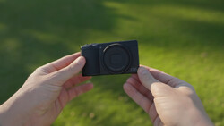 Hands holding a compact black mirrorless camera body against a blurred green outdoor background.