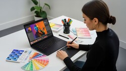 Designer working on color grading at laptop with paint swatches and color palettes on desk.