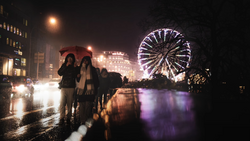 Wet city street at night with illuminated Ferris wheel reflecting in water and pedestrians in foreground.