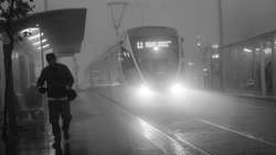 Runner in dark clothing passing an illuminated tram on a wet platform shrouded in fog.
