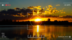 Camera viewfinder displaying a golden sunset reflected in still water with exposure and white balance controls visible.
