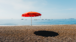 Red beach umbrella casting a dark shadow on sand, with calm ocean and moored boats beyond.