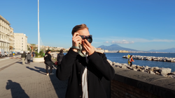 Person in black blazer and sunglasses holding camera to face on waterfront promenade.
