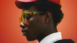 Profile portrait of a person wearing yellow-tinted sunglasses, red cap, and white collar against warm orange background.