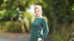 Woman in teal cardigan standing on a tree-lined path, looking toward camera.