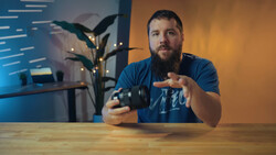 Bearded man in blue shirt demonstrating camera controls at wooden desk with warm backlighting.