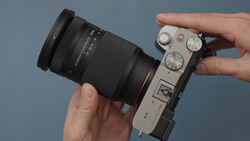 Hands holding a mirrorless camera with attached telephoto lens against a neutral blue background.
