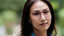 Woman wearing rose-gold rimmed glasses and blue shirt, smiling slightly at camera with soft natural outdoor lighting.