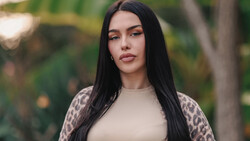 Woman with long black hair wearing a cream-colored top and leopard-print cardigan, photographed outdoors with blurred green foliage background.