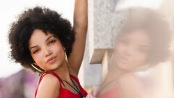 Woman with natural curly hair wearing red sleeveless top, arm raised, looking at camera with soft expression.