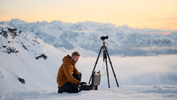 Photographer in brown jacket sitting in snow with camera mounted on tripod, mountain range and cloud inversion visible in background.