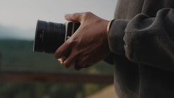 Photographer holding a telephoto lens while looking toward a blurred green landscape in overcast daylight.