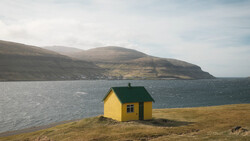 Bright yellow house with green roof on barren coastline beside dark ocean and distant mountains.
