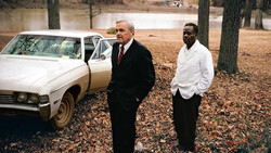 Two men in business attire standing beside a white sedan in a wooded area with a pond visible in the background.