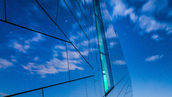 Modern glass building facade reflecting blue sky and clouds with geometric lines and reflections.