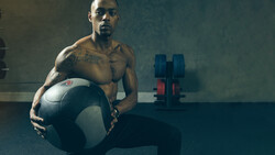 Shirtless man holding a medicine ball in a gym with dumbbells visible in the background.