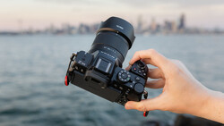 Hand holding a mirrorless camera with attached telephoto lens over water with city skyline in background.