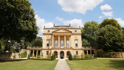 Neoclassical stone mansion with columned portico, symmetrical facade, and manicured grounds under blue sky.