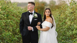 Bride and groom walking together through a vineyard in formal wedding attire.