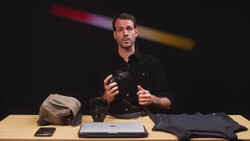 Man in black shirt gesturing while speaking at table with photography gear and laptop.