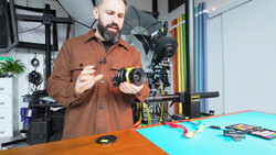 Bearded man in brown shirt holding a cinema camera lens in a studio workspace.