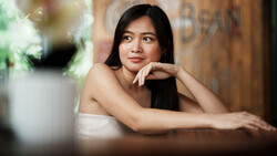 Woman with long dark hair resting chin on hand, looking toward camera in indoor setting with warm lighting.
