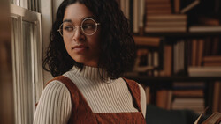 Portrait of a person wearing round glasses and a cream ribbed turtleneck with a rust-colored vest, positioned against bookshelves.