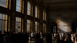 Crowds of visitors walking through a grand hall with tall arched windows casting golden light across the space.