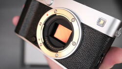 Mirrorless camera with exposed sensor and open body cap, held in hands against neutral background.