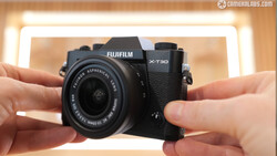 Hands holding a Fujifilm X-T30 mirrorless camera with attached lens against a blurred warm-toned background.