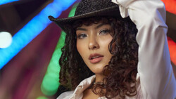 Woman wearing a black cowboy hat and white shirt against colorful neon-lit background.