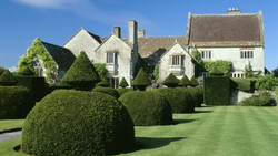 English country manor with manicured topiary hedges and lawn in foreground under clear blue sky.
