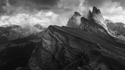 Dramatic black and white alpine landscape with jagged rock formations and distant snow-capped peaks beneath turbulent clouds.