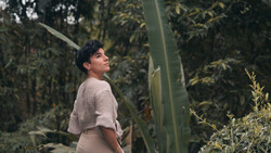 Person in beige shirt standing among tall green plants and cacti in a greenhouse environment.