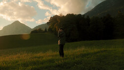 Person standing in a grassy meadow with forested mountains in the background during golden hour.