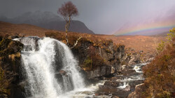 Waterfall cascading over dark rocks with rainbow arcing across misty valley landscape.