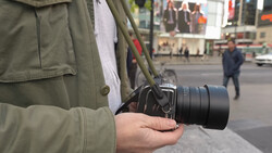 Photographer holding a mirrorless camera with telephoto lens on a city street.