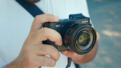 Photographer holding a Sony RX1 compact camera with Zeiss lens against a blue sky background.