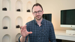 Man wearing glasses and plaid shirt gestures while speaking in a studio setting.