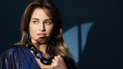 Woman with long brown hair wearing a blue sequined jacket and black chain necklace against a dark blue background.
