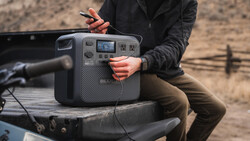 Person holding a portable power station with digital display while sitting in truck bed outdoors.
